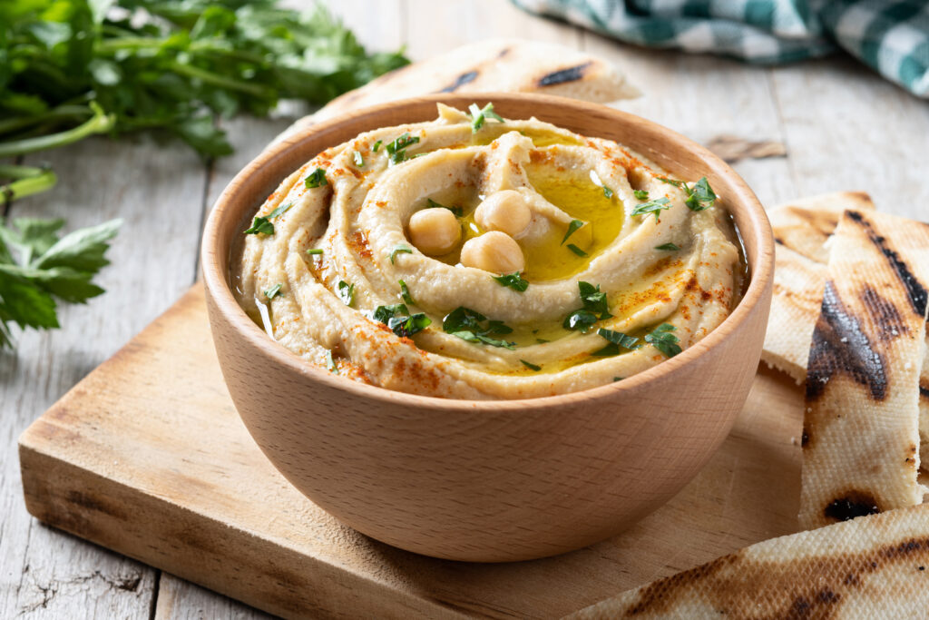 Imagen de un tazón de hummus de garbanzos casero, adornado con un chorrito de aceite de oliva, pimentón y perejil fresco, acompañado de bastones de zanahoria y pan pita.