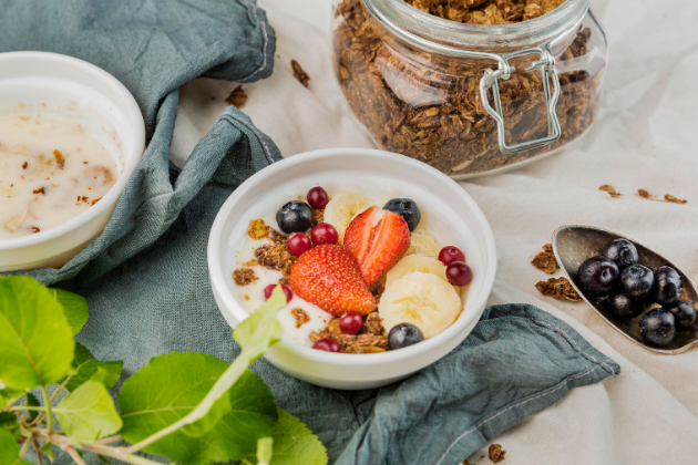 Bol de yogur con granola, fresas, arándanos, plátano y grosellas sobre una mesa, representando un snack saludable para el trabajo o la escuela.