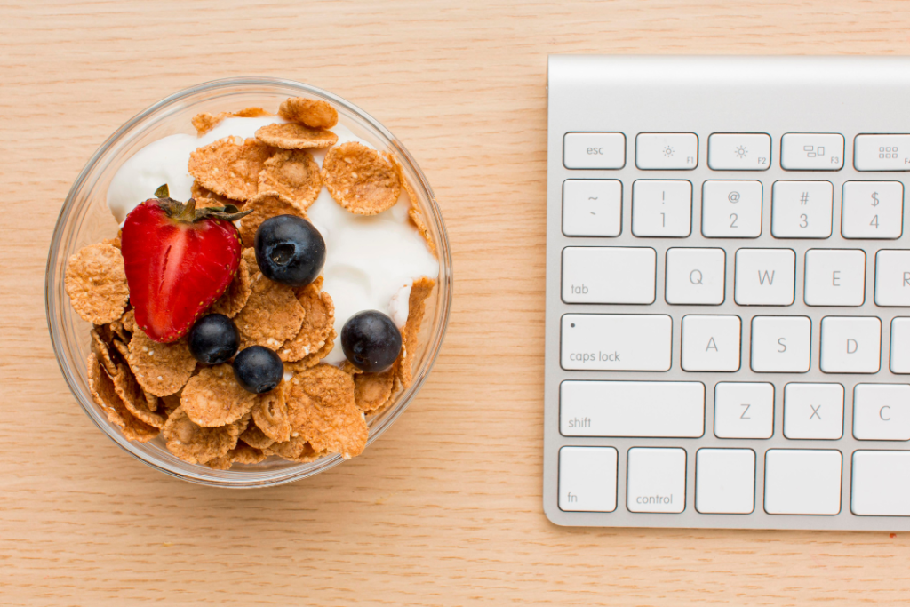 Bol de cereales integrales con yogur, fresas y arándanos junto a un teclado de ordenador, representando un snack saludable para el trabajo o la escuela.