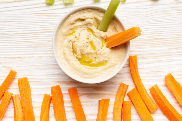 Bol de hummus con aceite de oliva acompañado de palitos de apio y zanahoria sobre una mesa de madera clara, representando un snack saludable y rápido.